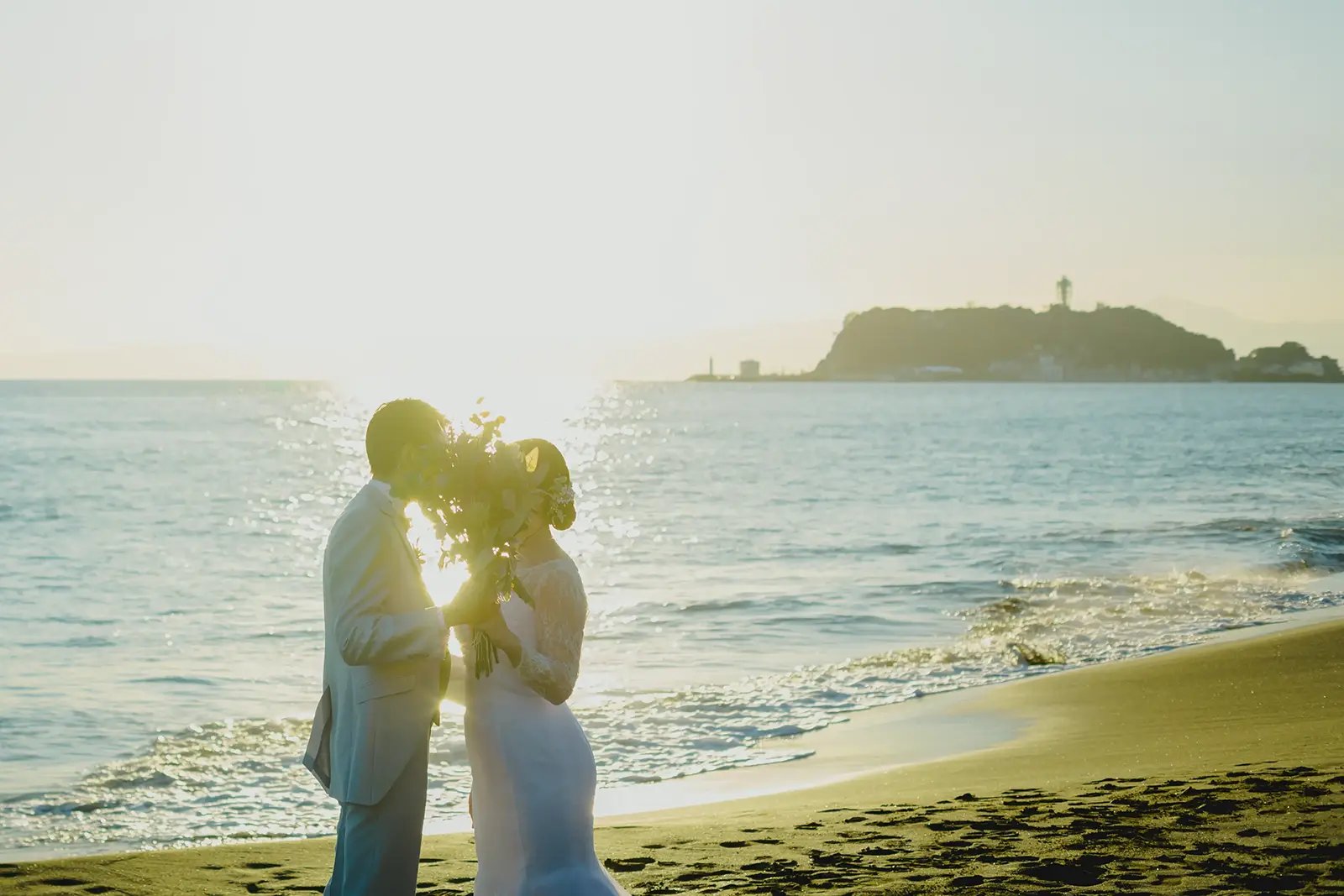 神奈川県・一色海岸の砂浜にて、夕日をバックにブーケを持って見つめ合う新郎新婦のシルエット写真。穏やかな波打ち際での幻想的なウェディングフォト。