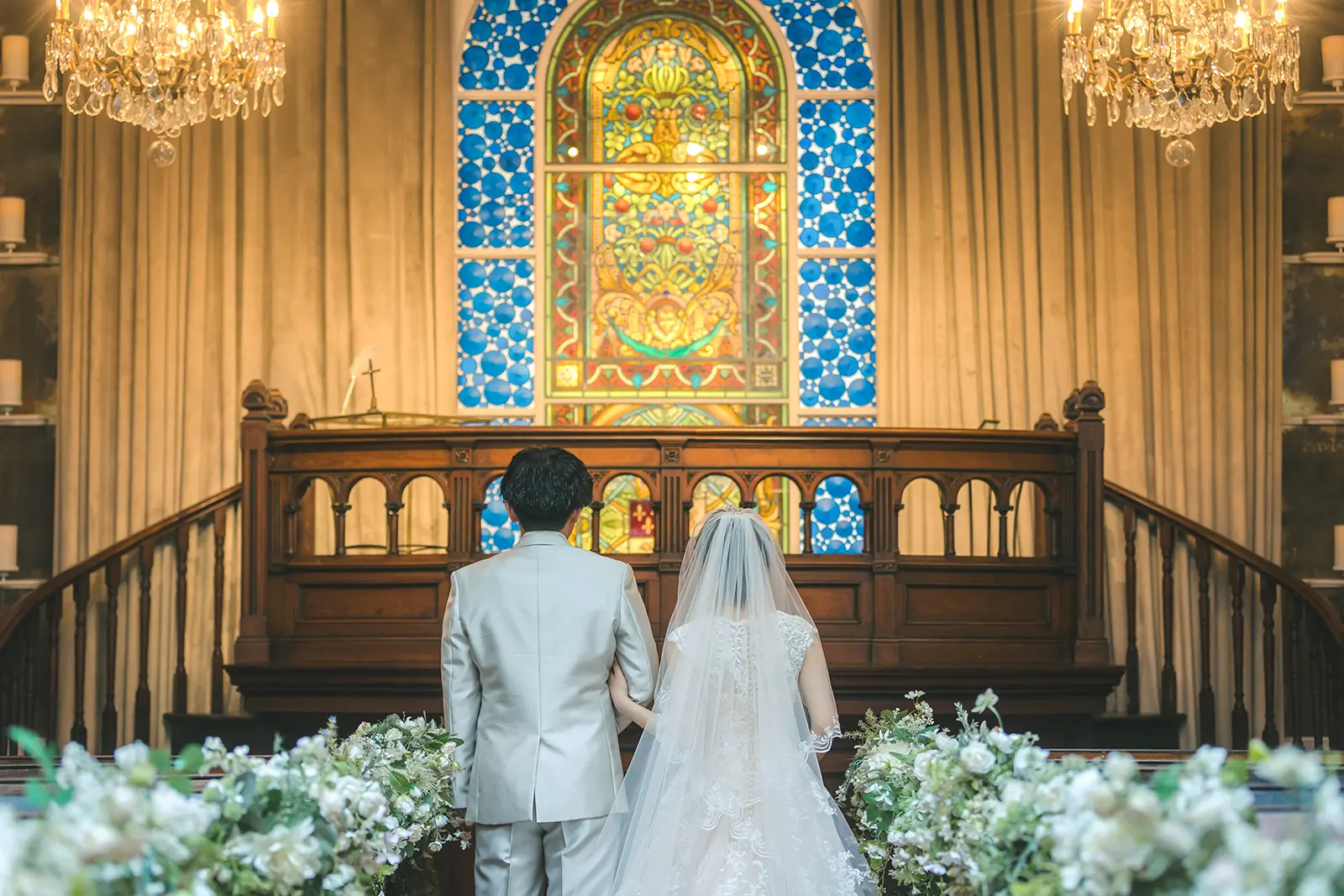 横浜のチャペルにて、美しいステンドグラスを背景に立つ新郎新婦の後ろ姿。アンティークな木の調度品と柔らかな光に包まれた、厳かなチャペルフォトウェディング。