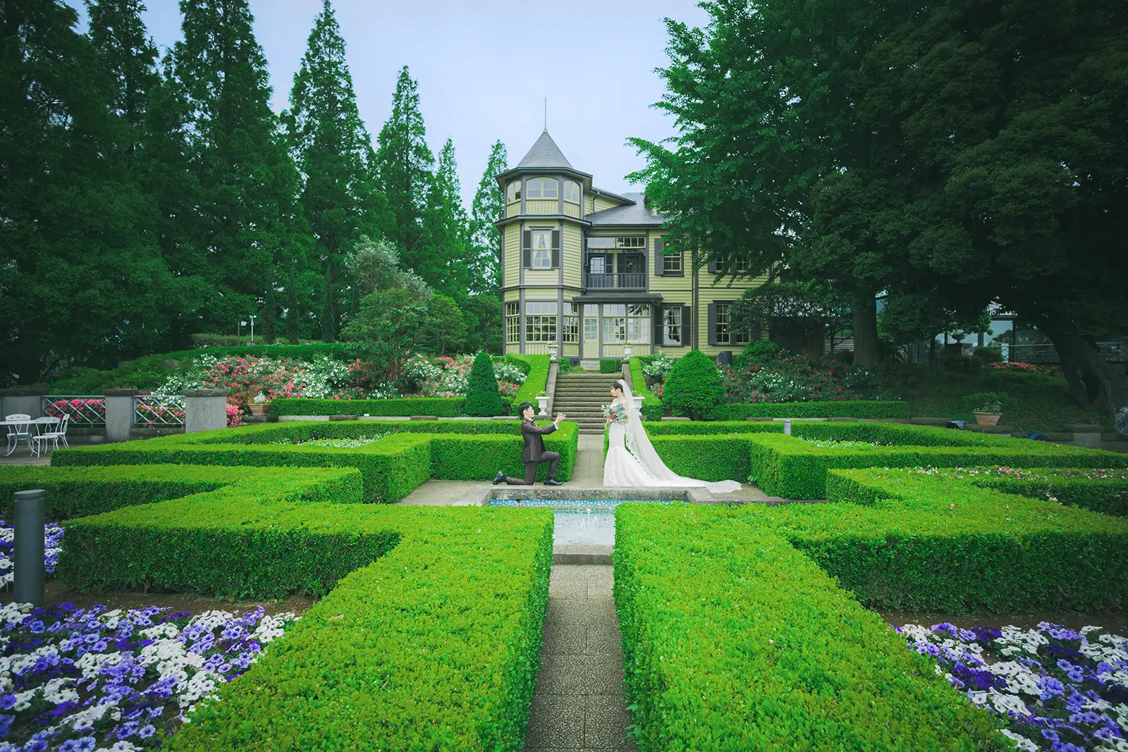 横浜・イタリア山庭園の幾何学的な迷路と緑色の洋館を背景に、膝をついてプロポーズする新郎と微笑む新婦のドラマチックなフォトウェディング。フォトジェリッシュによる特別ロケーション前撮り。
