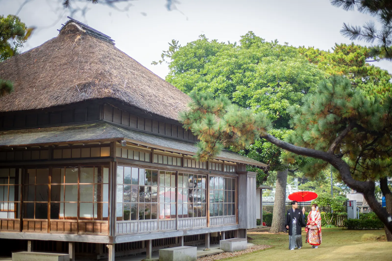 横浜・旧伊藤博文金沢別邸の茅葺き屋根の古民家を背景に、紋付袴と色打掛姿の新郎新婦が赤い和傘を差して歩く、庭園での和装フォトウェディング。