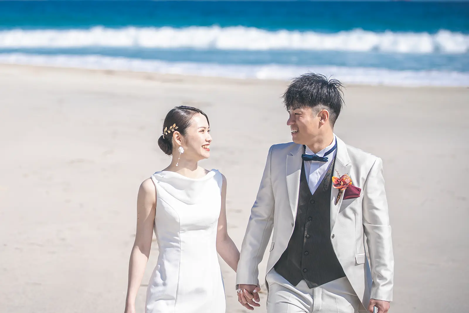 伊豆・白浜の砂浜にて、手をつないで笑顔で歩くウェディングドレスとタキシード姿の新郎新婦。青い海を背景にした明るく爽やかな洋装フォトウェディング。