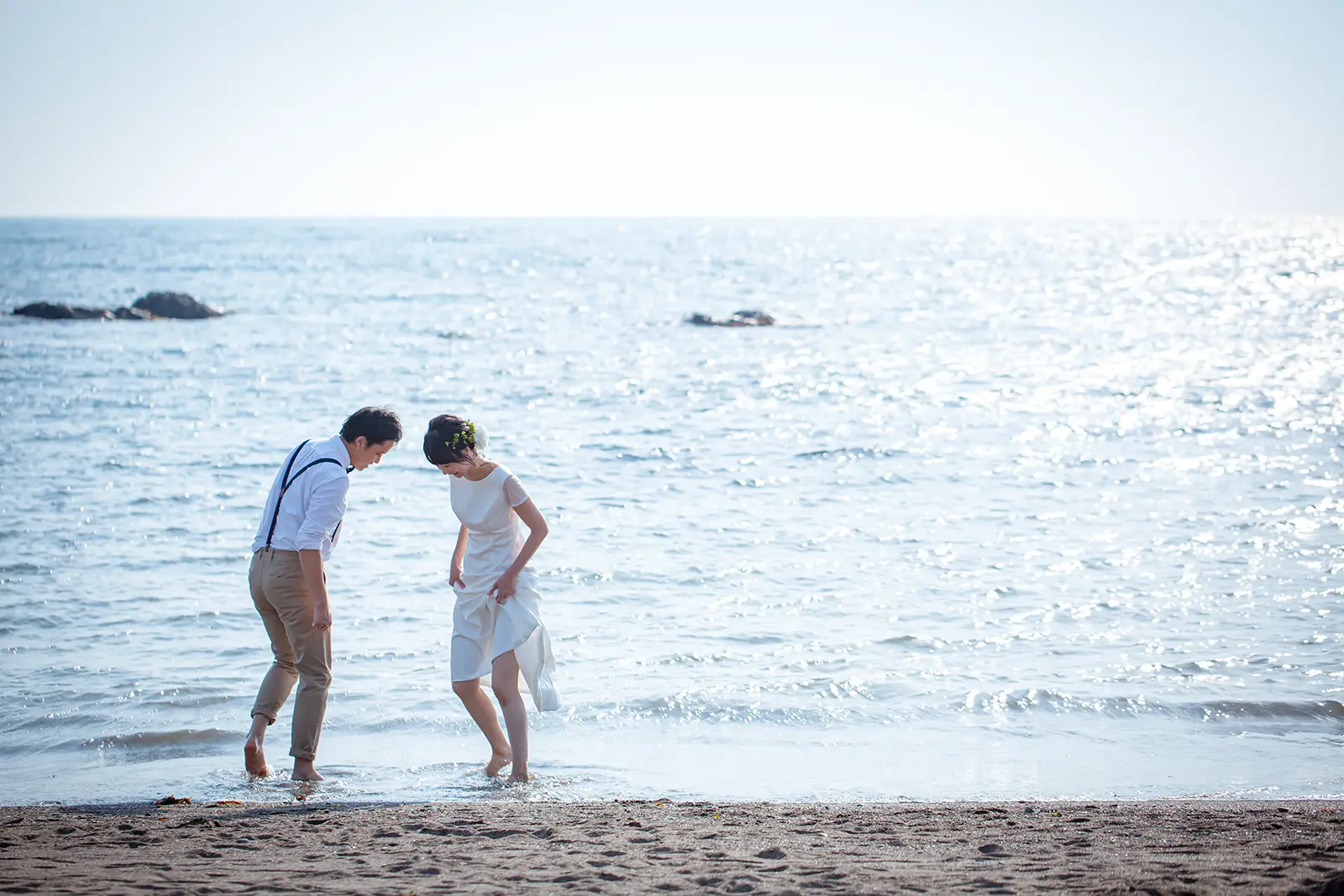 金沢の海岸にて、波打ち際で寄り添うウェディングドレスとタキシード姿のカップル。夕暮れ時や美しい海を背景にした、フォトジェリッシュによる洗練された前撮り風景。