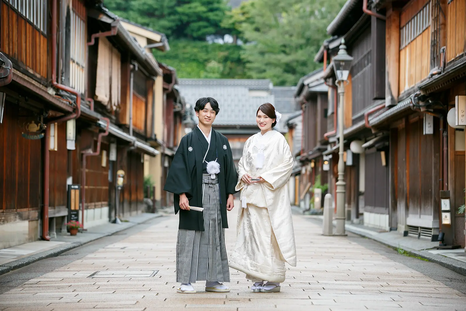 石畳の通りが続く金沢・ひがし茶屋街の情緒ある街並みを背景に、紋付袴と白無垢姿で佇む新郎新婦の全身フォト。伝統的な町家が並ぶ、格式高い和装前撮り。