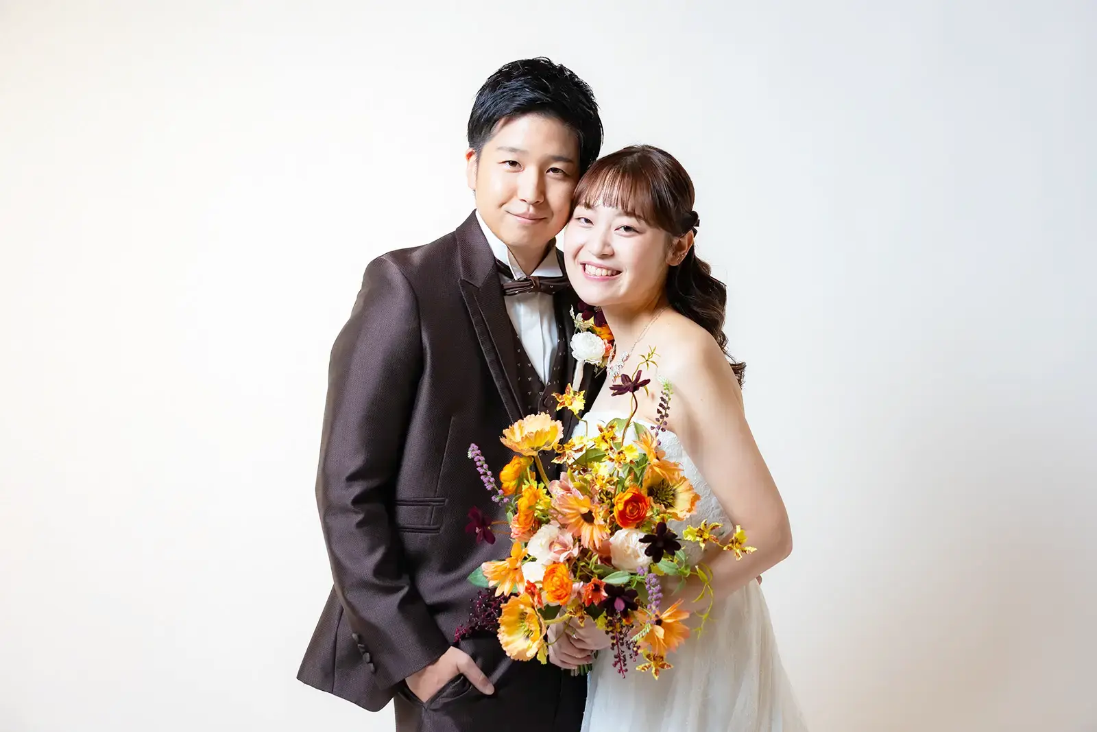シンプルな白背景のスタジオにて、ブラウンのタキシードを着た新郎と、ウェディングドレスを着て色鮮やかなオレンジ・黄色のブーケを持った新婦が、寄り添って満面の笑みを浮かべる洋装ウェディングフォト。