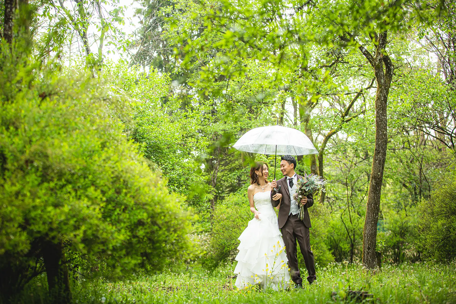 軽井沢の豊かな緑の中で、透明な傘をさして寄り添うウェディングドレス姿の花嫁とタキシード姿の新郎。フォトジェリッシュによる、雨の日ならではの幻想的で美しい前撮り風景。