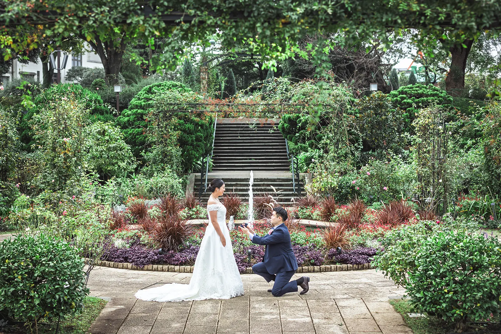 横浜・港の見える丘公園のイングリッシュローズガーデンにて、新緑のアーチと噴水を背景に、片膝をついてプロポーズをする新郎と微笑む新婦。ドラマチックなウェディングフォト。
