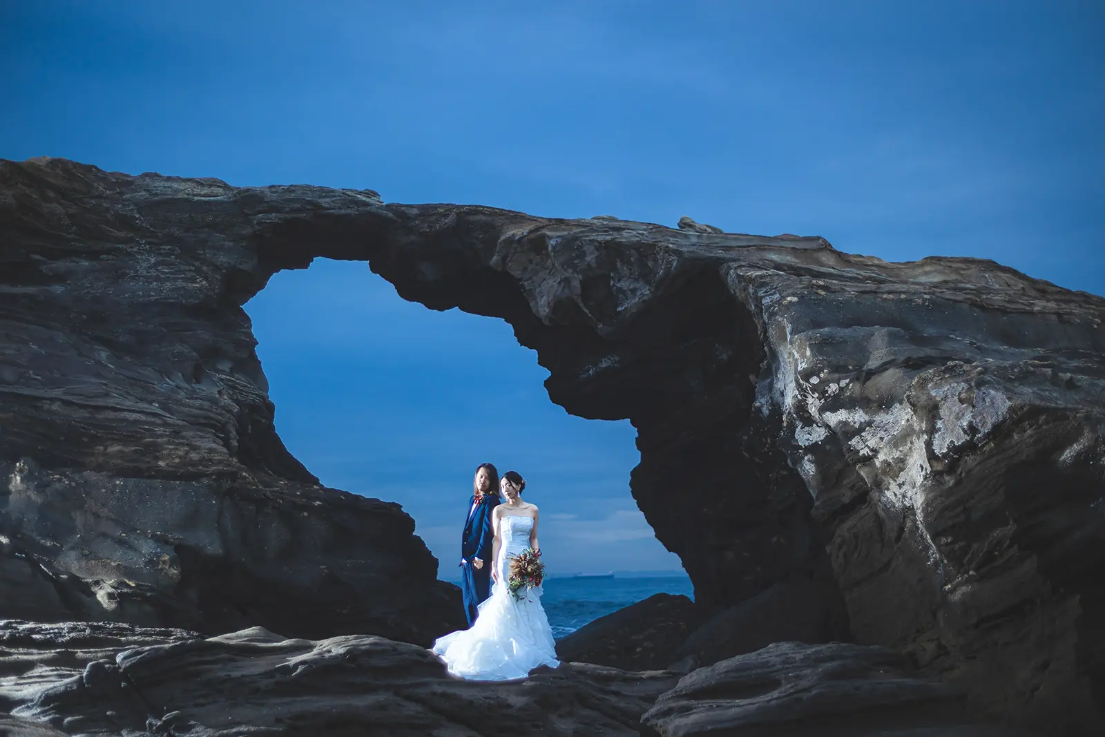 三浦・城ヶ島の断崖絶壁を背景に、ウェディングドレスとタキシード姿で佇むカップル。フォトジェリッシュによる、雄大な自然を活かしたドラマチックなフォトウェディング。