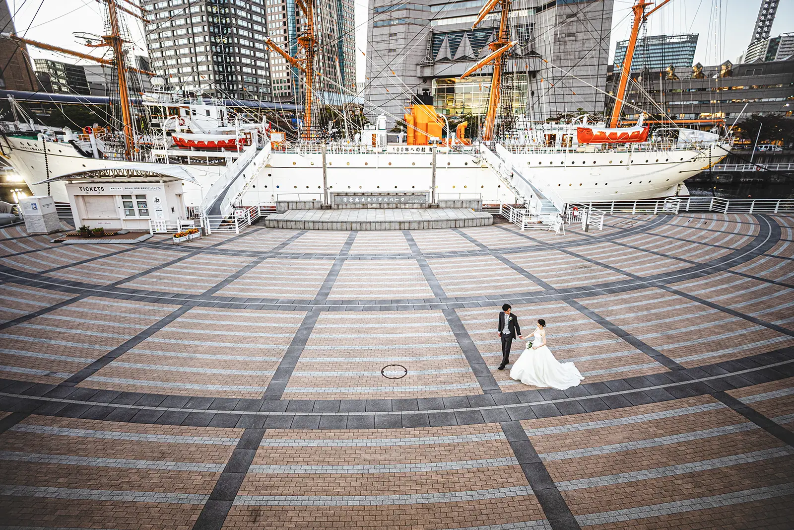 横浜・日本丸メモリアルパークの広場にて、帆船「日本丸」を背景に歩くウェディングドレス姿の新郎新婦。みなとみらいの近代的なビル群と帆船が調和するロケーションフォト。