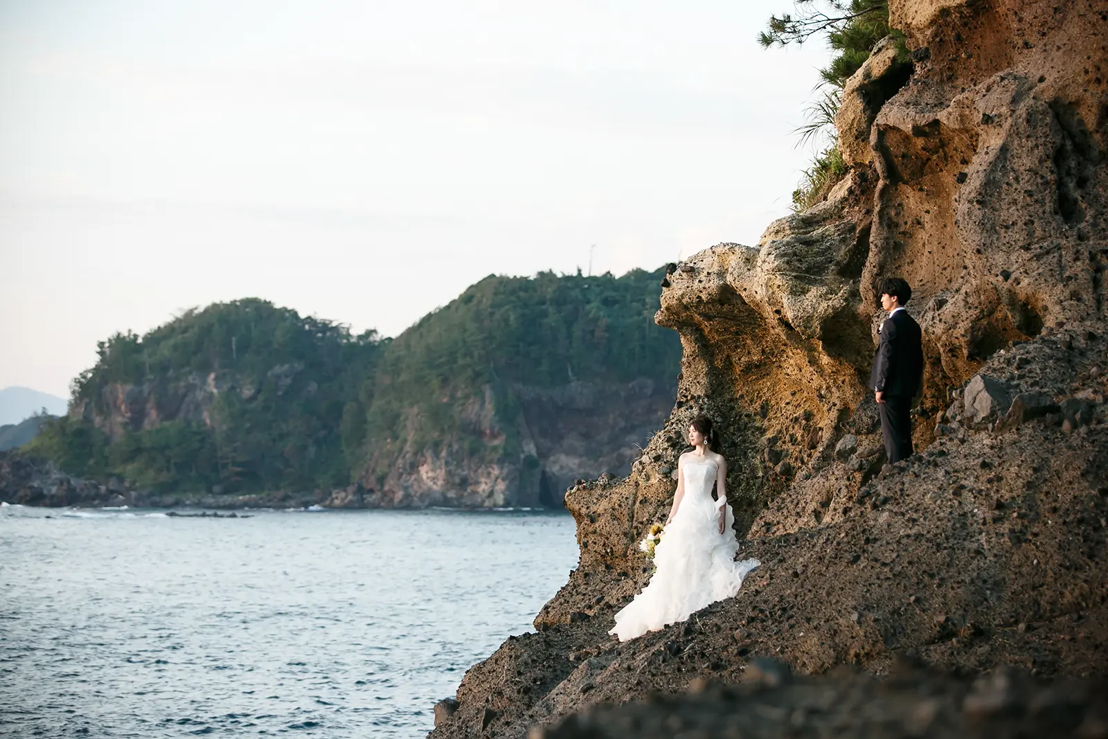 石川県・能登の景勝地「雁門」にて、日本海の波打ち際と奇岩を背景に立つ新郎新婦。壮大な自然が織りなす、ダイナミックで情緒あふれるロケーションフォト。