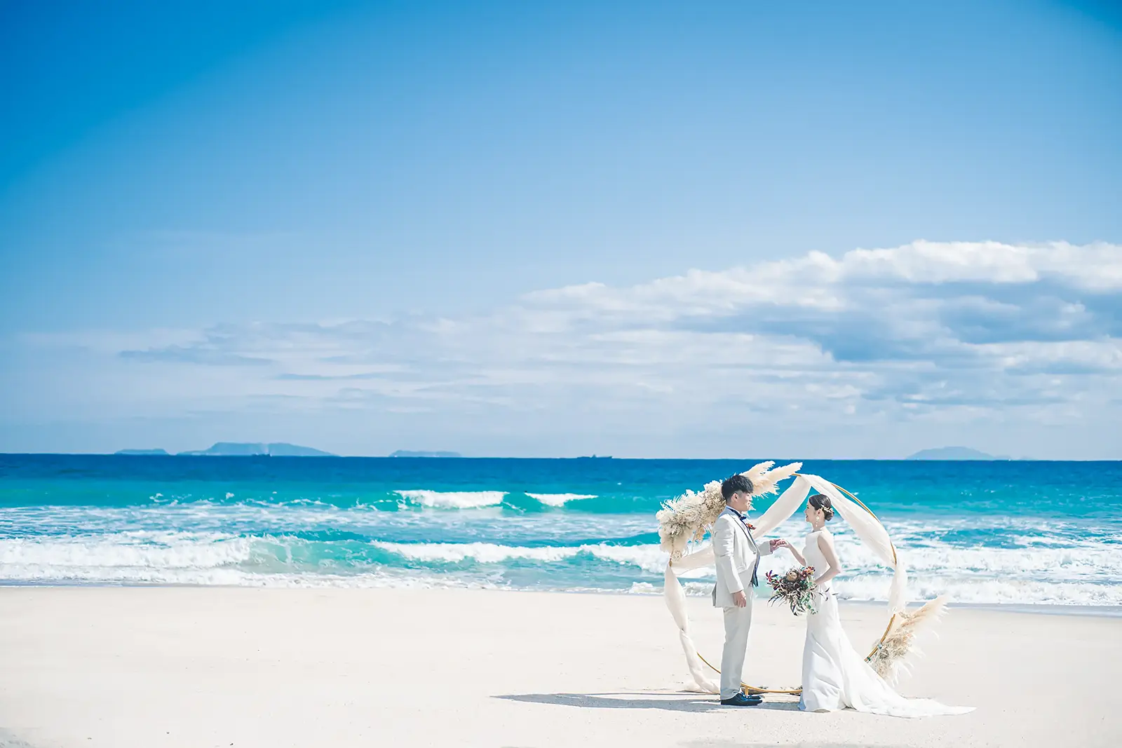 伊豆下田の美しい青い海と白い砂浜を背景に、ウェディングアーチの前で手を取り合う新郎新婦。開放感あふれるビーチでのフォトウェディング。