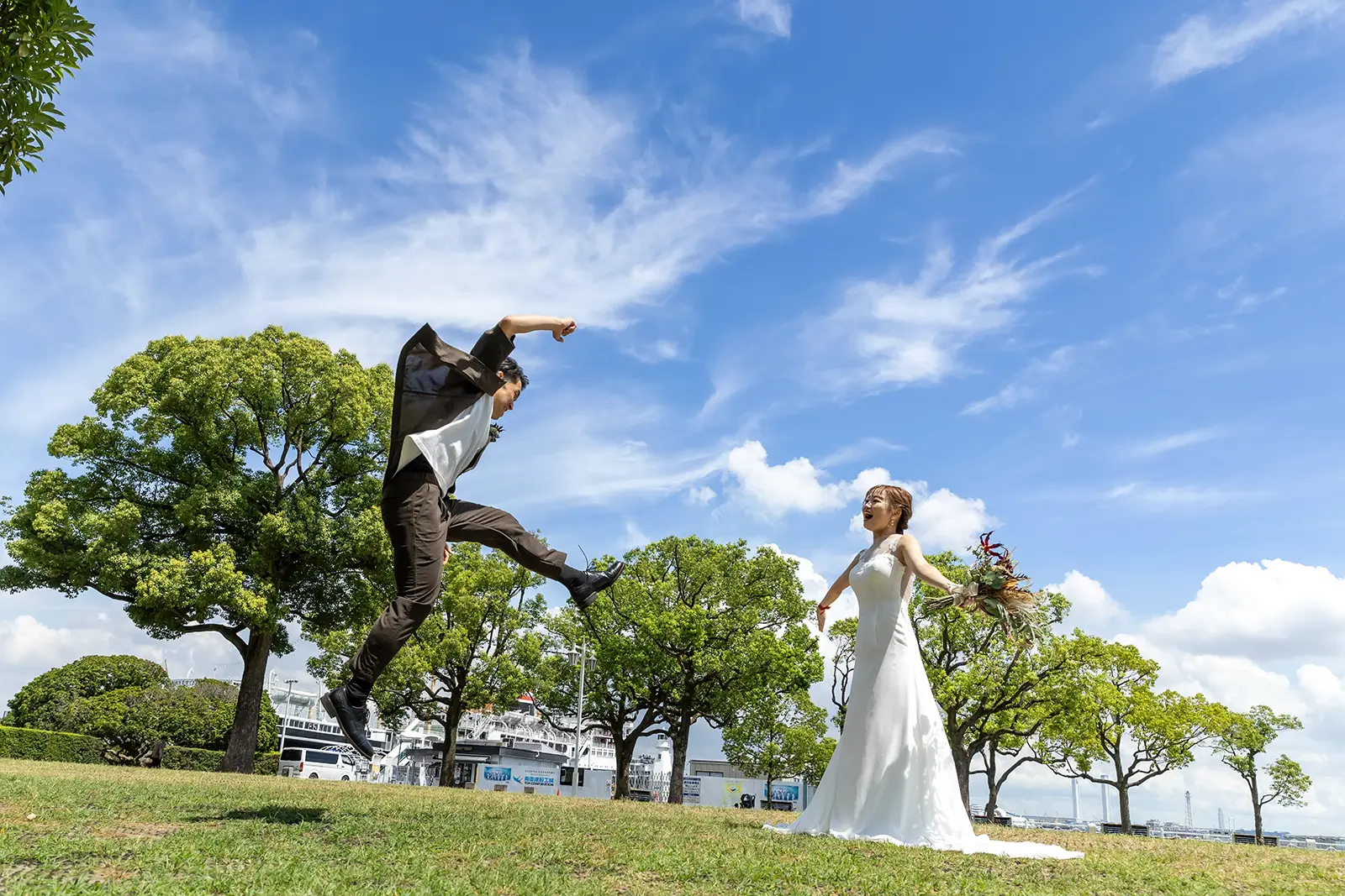 鮮やかな青空と緑の芝生をバックに、ウェディングドレス姿の女性に向かってジャンプするタキシード姿の男性。フォトジェリッシュによる、開放感あふれる楽しいフォトウェディングの一枚。