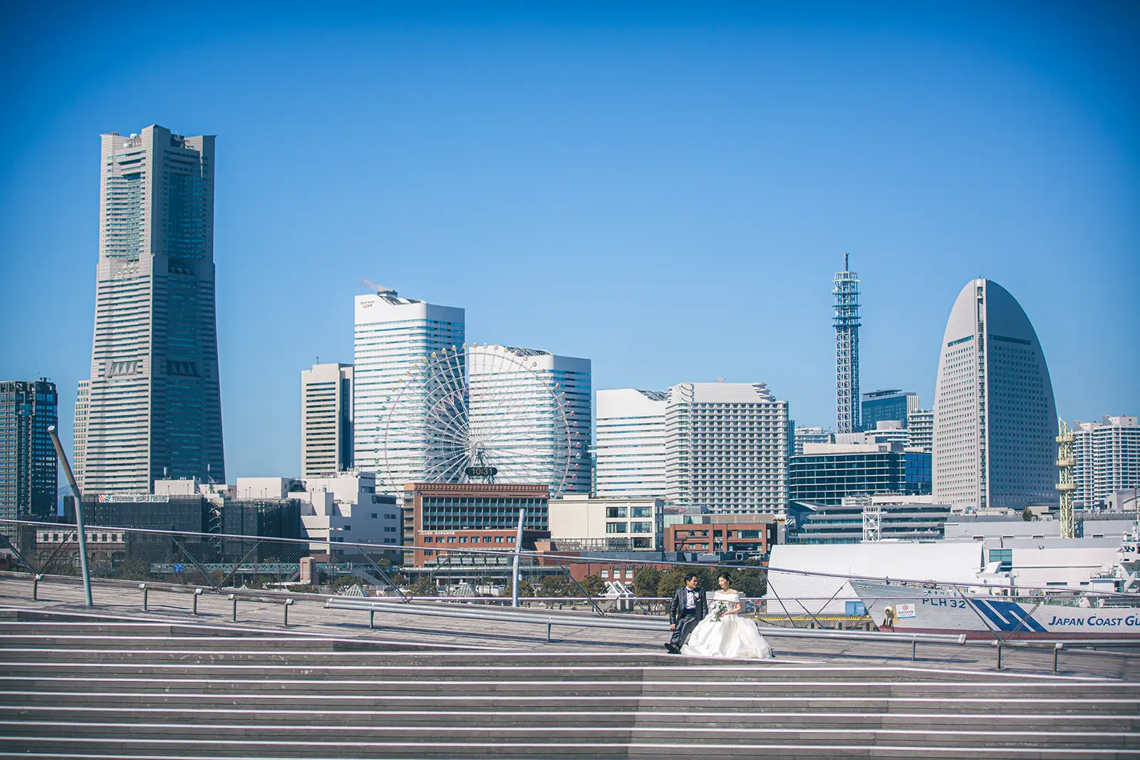 横浜大桟橋のウッドデッキで撮影された、みなとみらいのスカイラインを背景にしたカップルのフォトウェディング写真。フォトジェリッシュによる洗練された前撮り風景。