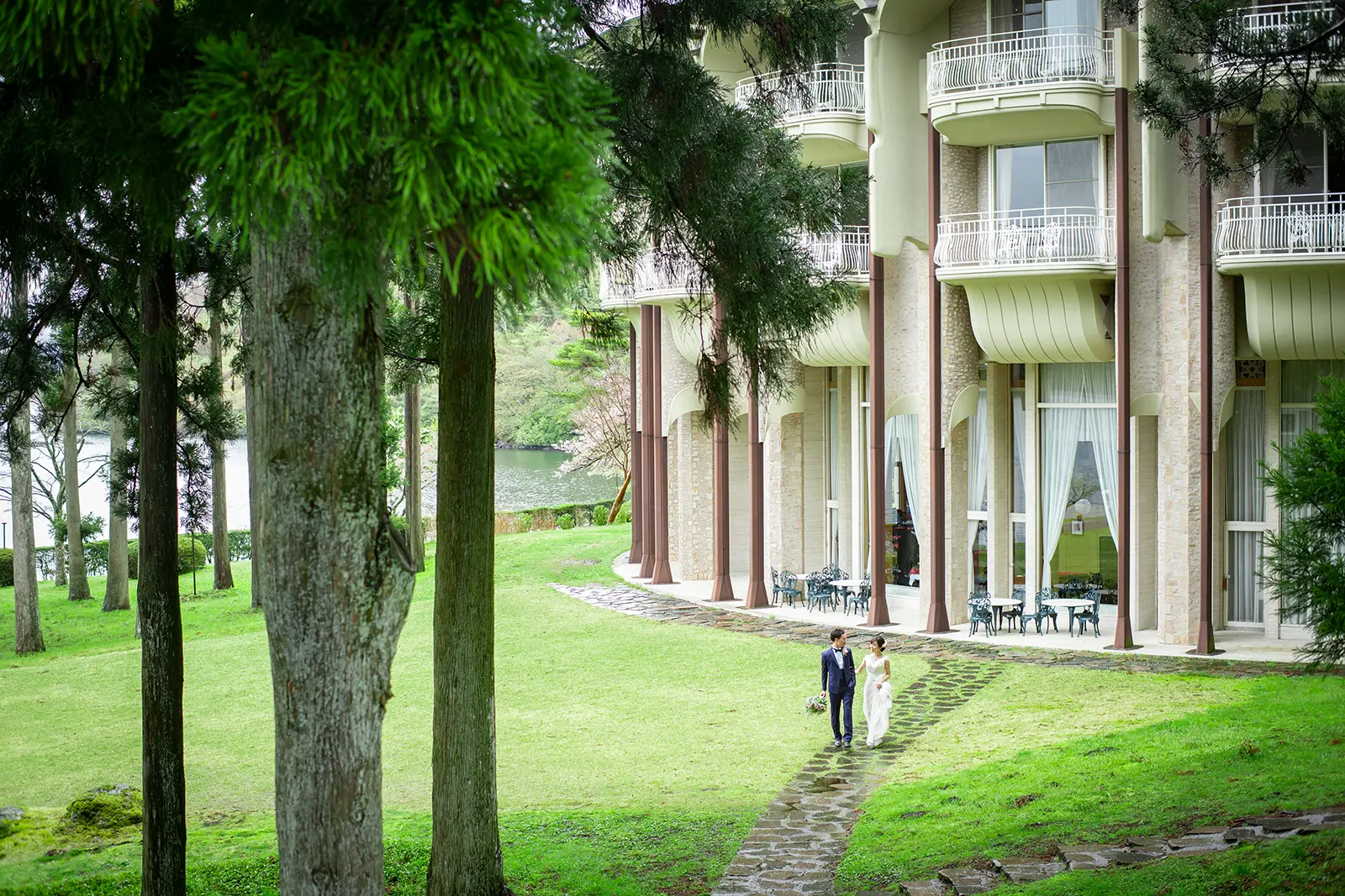箱根・芦ノ湖畔に佇むクラシックホテルのガーデンにて、新緑の森を背景に石畳を歩くウェディングドレスとタキシード姿の新郎新婦。厳かで洗練されたロケーションフォト。