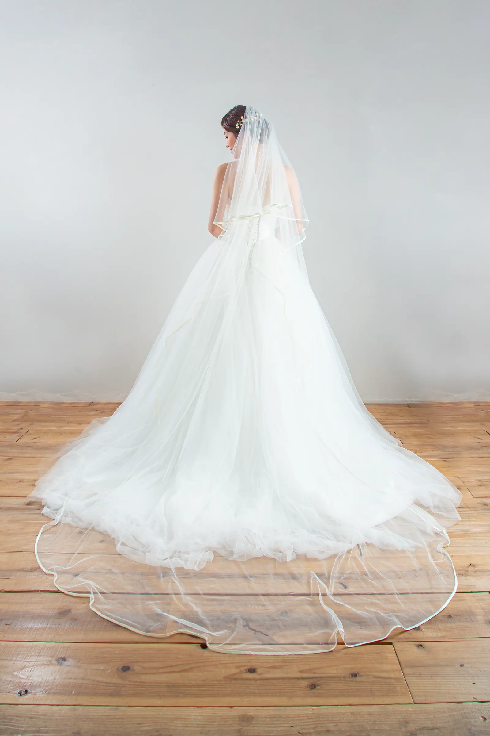 チャペルのバージンロードに美しく広がる、裾に繊細なレースをあしらったロングベールの全身後ろ姿。ウェディングドレスのバックスタイルを引き立てる一枚。