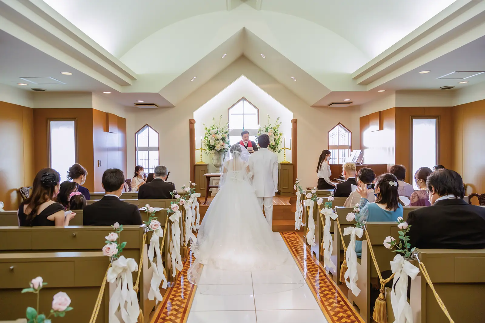 光が差し込むチャペルにて、祭壇に向かって立つウェディングドレス姿の新婦とタキシード姿の新郎の後ろ姿。両脇のベンチには家族が見守る、フォトジェリッシュの家族挙式風景。