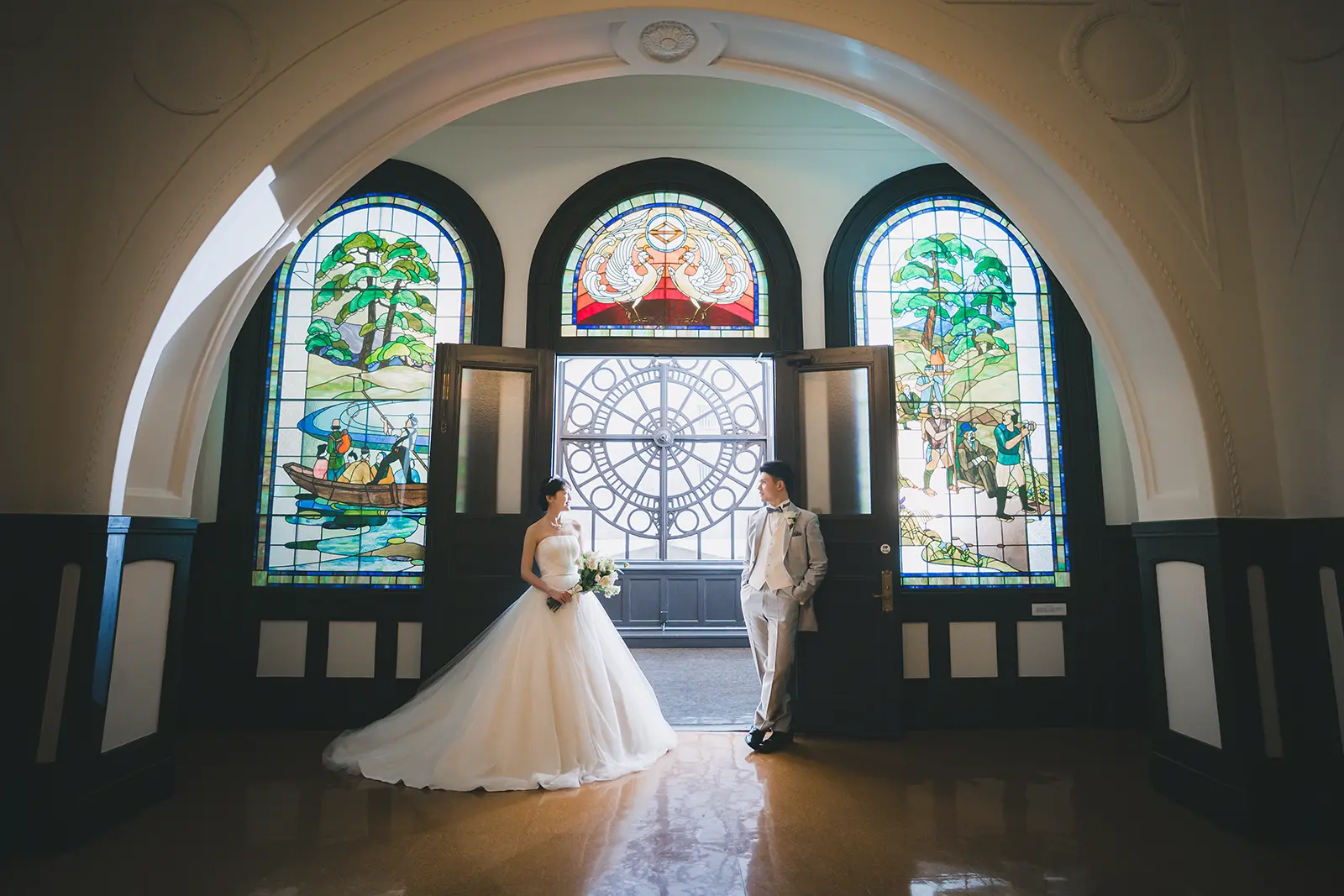 横浜市開港記念会館の美しいステンドグラスを背に立つ、ウェディングドレスとタキシード姿のカップル。フォトジェリッシュによるクラシカルな洋館前撮り。