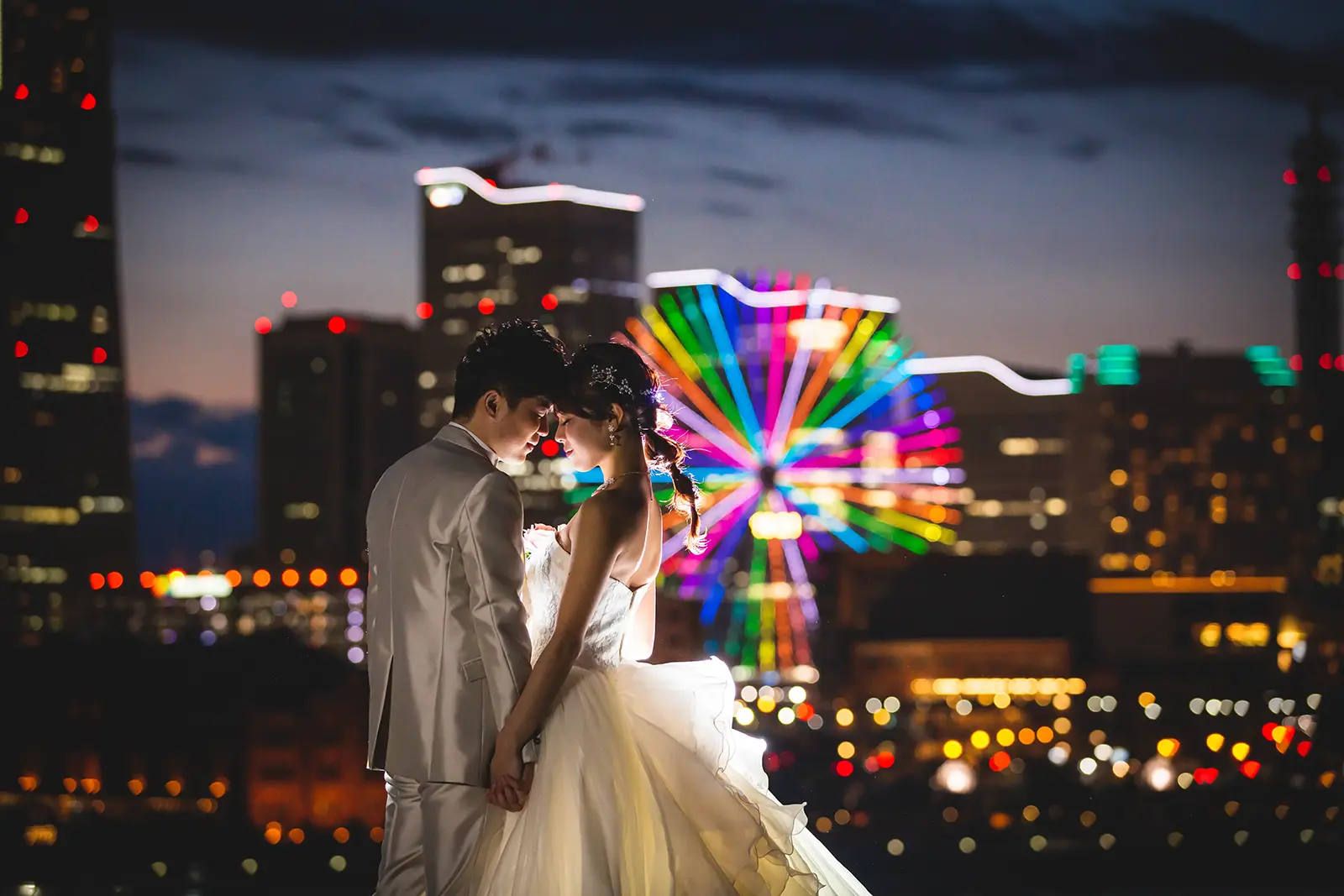 横浜・大さん橋にて、みなとみらいの夜景とライトアップされた観覧車を背景に見つめ合うウェディングドレスとタキシード姿のカップル。フォトジェリッシュによるドラマチックな夜景フォト。